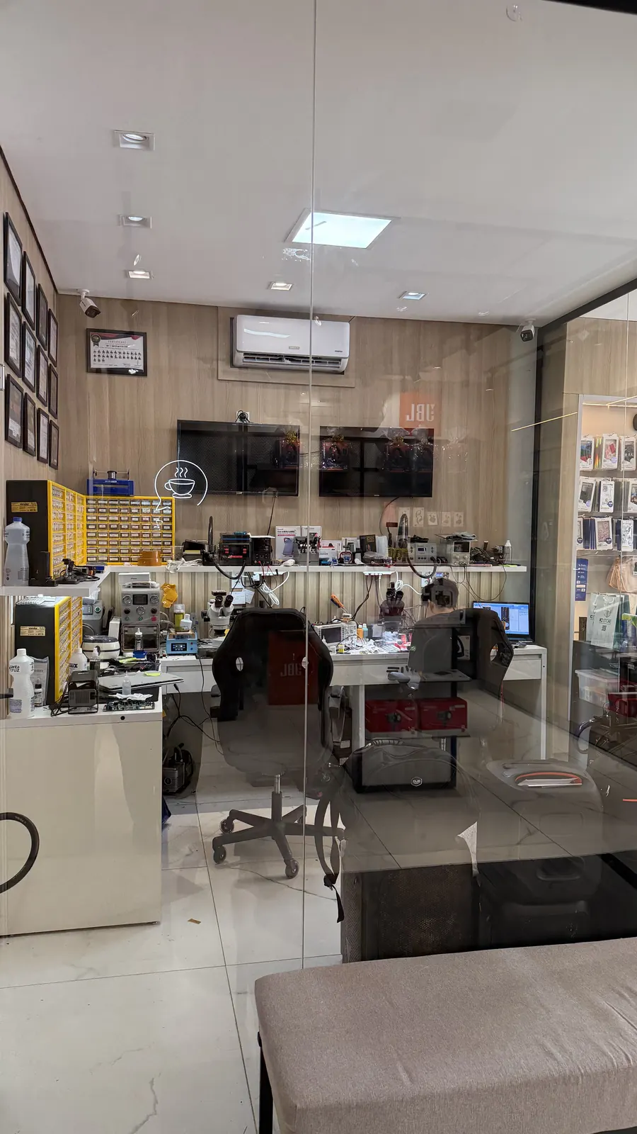 Bancada de reparos da IC Store vista pelo vidro: microscópios, estações de solda, gavetas de componentes e parede de certificados